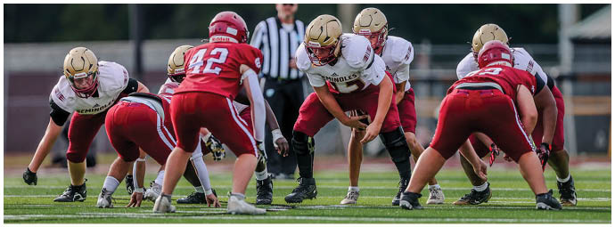 Two opposing American football teams in red and white uniforms are lined up on a field preparing for a play with a referee standing behind them. Description generated by AI