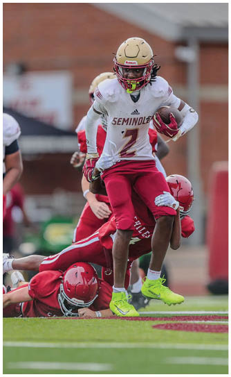 Two people in red football uniforms are on the ground while a person in a white and red football uniform with a gold helmet stands behind them on a football field. Description generated by AI