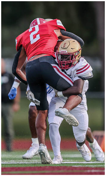 A person in a white football uniform tackles another person wearing a red jersey and black pants during a game on a field. Description generated by AI