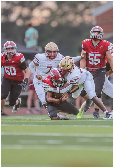 A group of people wearing football uniforms are on a field with one person in white tackling another person in red who is holding a football. Description generated by AI
