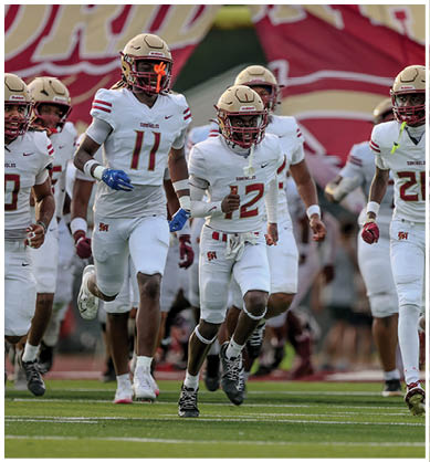 Two people in white football uniforms with helmets are running on a field during a game. Description generated by AI