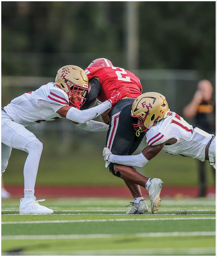 Three football players are engaged in a tackle on a grassy field, with two players in white uniforms and gold helmets tackling a player in a red jersey and black pants. Description generated by AI