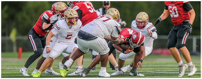 A group of people wearing football uniforms are engaged in a play on a grassy field with one person in red being tackled by several people in white and gold helmets. Description generated by AI