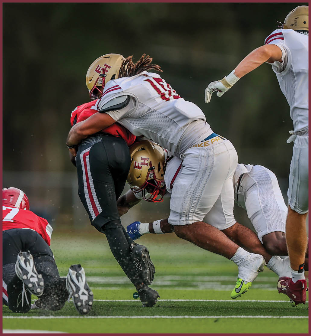 Two people in football gear are engaged in a tackle on a grassy field, with one wearing a gold helmet marked with “FH" and the other in black pants with red and white stripes. Description generated by AI