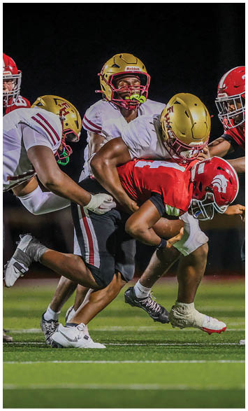 Four football players in helmets and uniforms are engaged in a tackle on a grassy field during a nighttime game. Description generated by AI