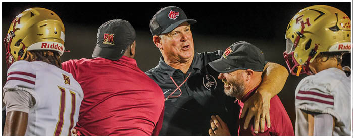 Four people wearing football helmets and two people in caps and sportswear are engaged in a lively conversation or celebration on a football field. Description generated by AI