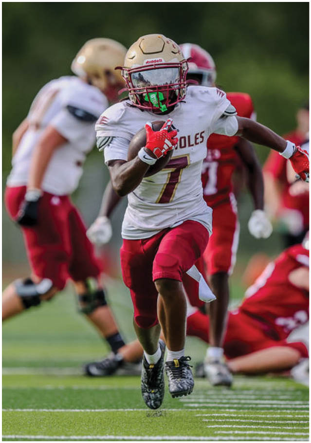 A person in a white and red football uniform is running with the ball on a green field while other players and a referee are visible in the background. Description generated by AI