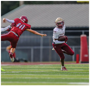 A person in a white and maroon football uniform runs with the ball while two people in red uniforms leap and reach out to try to tackle them on a football field. Description generated by AI