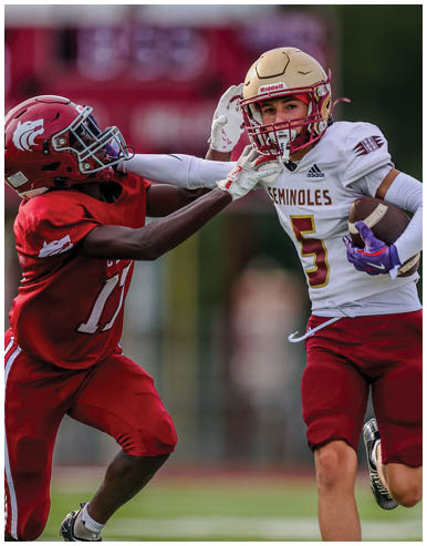 Two people in football uniforms are actively competing on a field, with one person running while holding the ball and the other reaching out to block or tackle. Description generated by AI