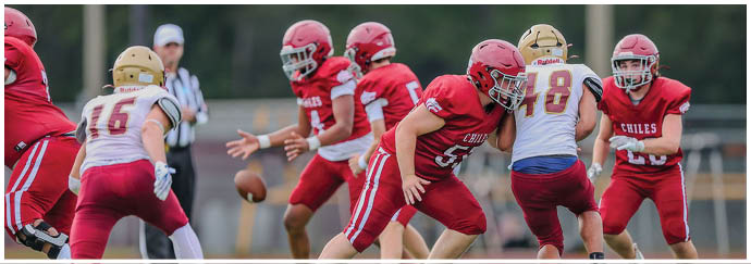 People wearing red and white football uniforms are actively engaged in a play on a football field with one person about to catch a football in midair. Description generated by AI