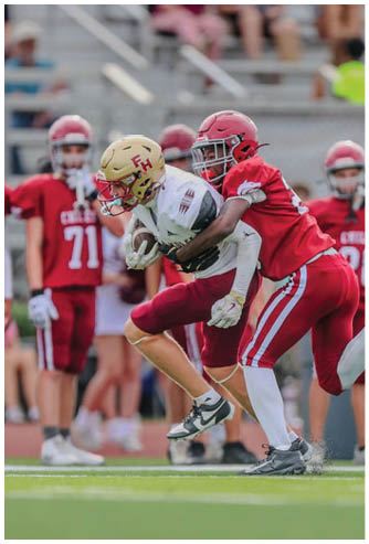Two people in football uniforms are engaged in a tackle on a grassy field with others in the background. Description generated by AI