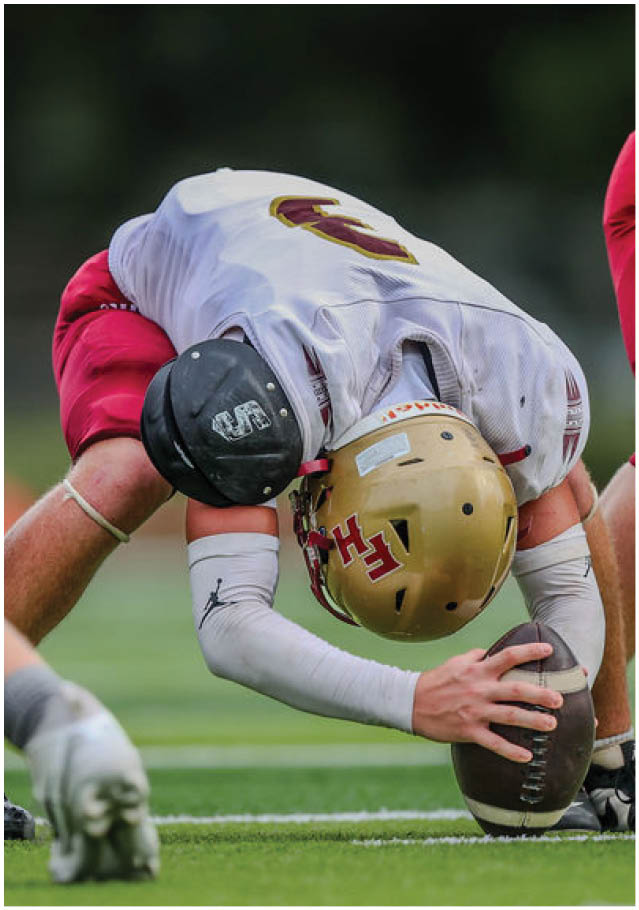 A person in football gear is crouched down holding a football on a field while other players in red and white uniforms are positioned nearby. Description generated by AI
