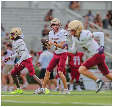 Three people in football uniforms are on a field with one holding a football and preparing to throw while the others are running. Description generated by AI