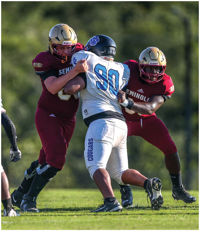 People in football uniforms are engaged in a play on a grassy field with one person in a maroon uniform holding the ball while two others in white uniforms attempt to block or tackle. Description generated by AI