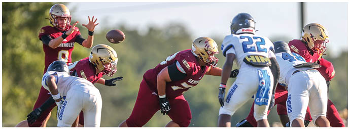 Adults in football uniforms are positioned on a field with one player in a maroon uniform throwing a football while others in maroon and white uniforms prepare for the play. Description generated by AI