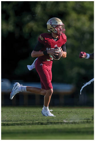 A person wearing a maroon football uniform and gold helmet is running on a grassy field while holding a football. Description generated by AI