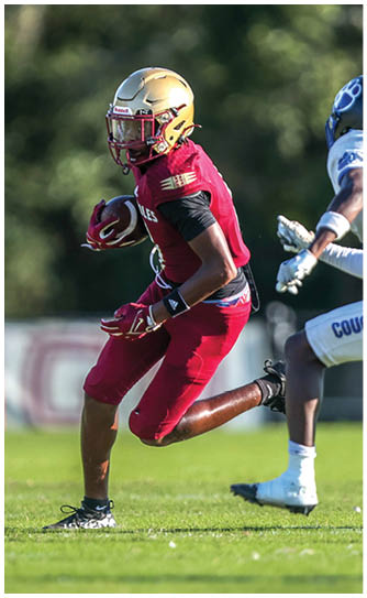 A person in a red football uniform and gold helmet is running with the ball while another person in a white uniform with “COUGARS" on the pants is attempting to catch up on a grassy field. Description generated by AI