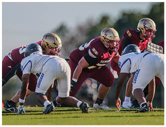 Four football players in helmets and uniforms are positioned on a grassy field facing each other, preparing for a play. Description generated by AI