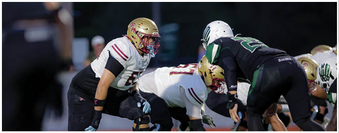 People wearing football helmets and uniforms are positioned on a field, appearing ready for a play during a game. Description generated by AI
