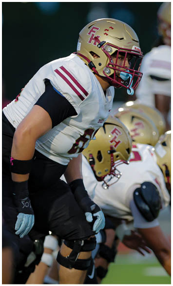 A person in a football uniform with a gold helmet and white jersey is crouched in a ready position on a football field with teammates lined up behind. Description generated by AI