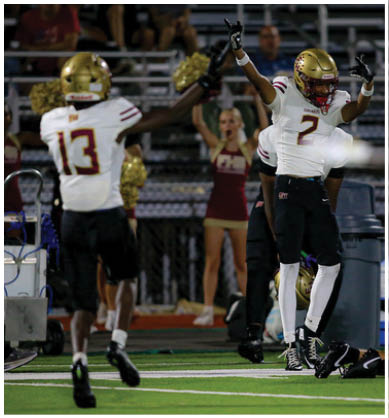 Four football players in white jerseys and gold helmets are lined up on a field with one player pointing forward under stadium lights. Description generated by AI