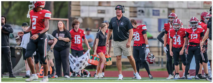 People in sports uniforms and casual clothing stand on the sidelines of a football field during a game or practice. Description generated by AI