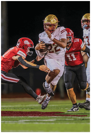 A person in a white football uniform with a gold helmet is running with the ball while two people in red and black football uniforms attempt to tackle them on a nighttime field. Description generated by AI