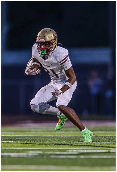 A person in a white football uniform with a gold helmet is running on a field while holding a football during a nighttime game. Description generated by AI