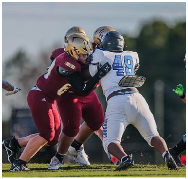 Two people in maroon football uniforms are tackling a person in a white football uniform during a game on a grassy field. Description generated by AI