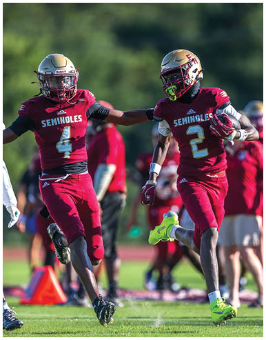 Two football players in red Seminoles uniforms are running on a field with one player holding a football and the other reaching out to him. Description generated by AI