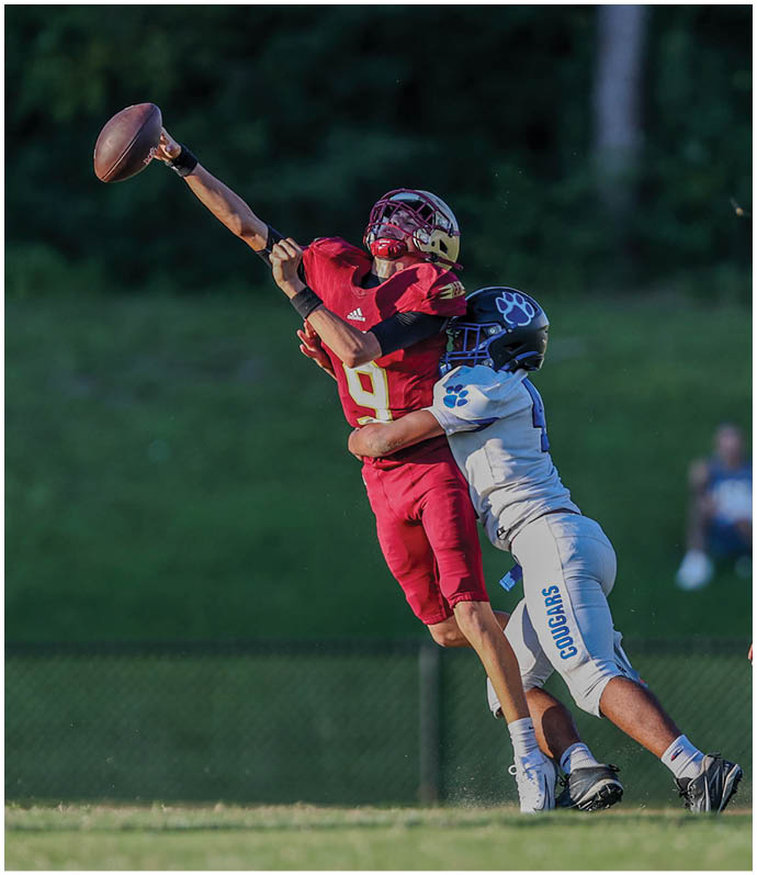 A person in red football gear is jumping and reaching out to catch a football while being closely defended by a person in white football gear on a grassy field. Description generated by AI