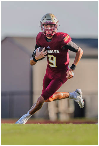 A person wearing a maroon football uniform with the number 9 is running on a field while holding a football. Description generated by AI