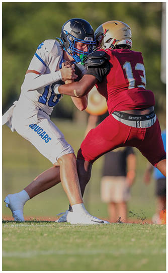 Two people in football uniforms are engaged in a play on a grassy field with one person wearing white and blue and the other wearing red and gold. Description generated by AI