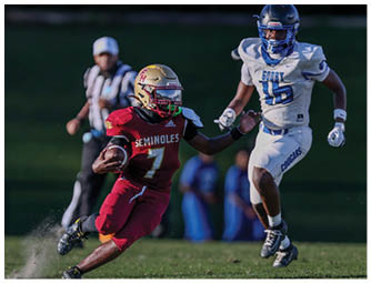 A person in a red football uniform is running on a field while two people in white and blue football uniforms are approaching from either side. Description generated by AI