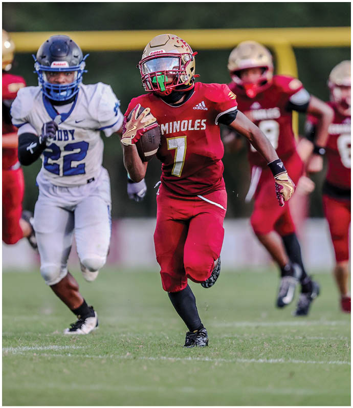 A person in a red football uniform with the number seven is running with the ball while being chased by a person in a white and blue football uniform on a grassy field. Description generated by AI