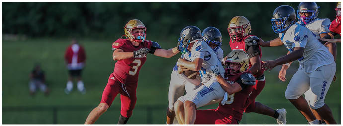 People wearing red and white football uniforms are engaged in a tackle on a grassy field during a game. Description generated by AI