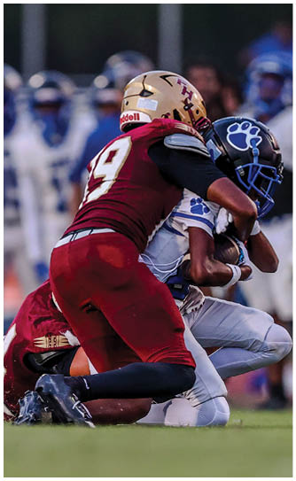 Two people in football uniforms are engaged in a tackle on a grassy field with other players and a coach blurred in the background. Description generated by AI