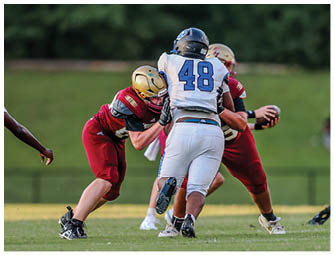 Two teams of people in football uniforms are engaged in a play on a grassy field with one person holding the ball and others blocking or tackling. Description generated by AI