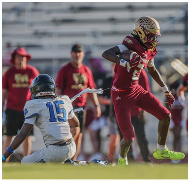 A person in a red football uniform is running with the ball while another person in a white uniform is sitting on the ground during a game on a field with blurred spectators in the background. Description generated by AI