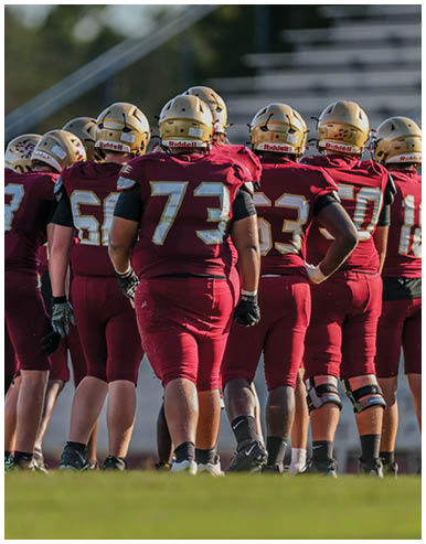 A group of people wearing maroon football uniforms and gold helmets stand together on a grassy field with empty bleachers in the background. Description generated by AI