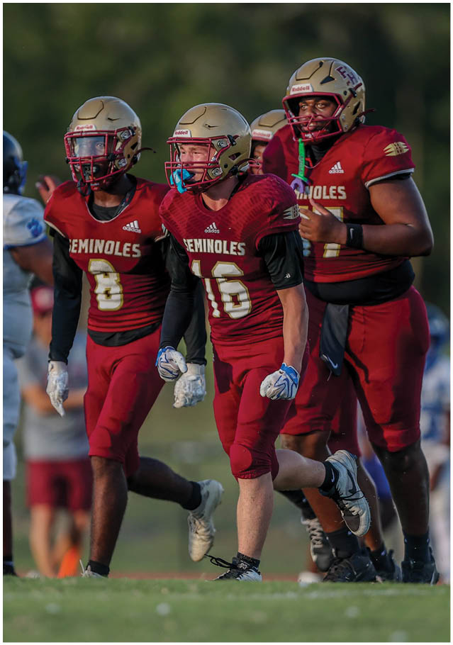 Three people wearing maroon football uniforms and helmets are running on a grassy field with blurred spectators in the background. Description generated by AI