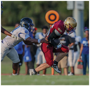 A person in a red football uniform is running with the ball while another person in a white and blue uniform attempts to tackle them on a grassy field. Description generated by AI