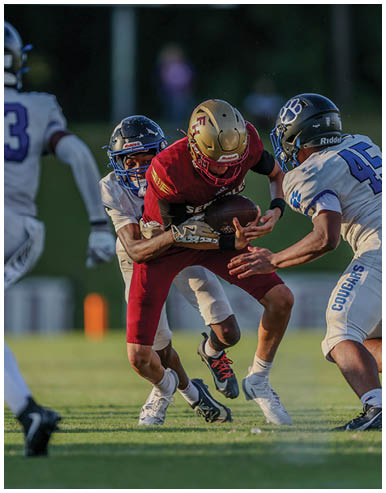 A person in a red football uniform is holding a football while being tackled by two people in white uniforms on a grassy field during a game. Description generated by AI