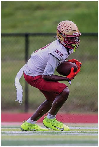A person wearing a football helmet, white jersey, red shorts, and neon yellow cleats is crouched on a football field holding a football. Description generated by AI