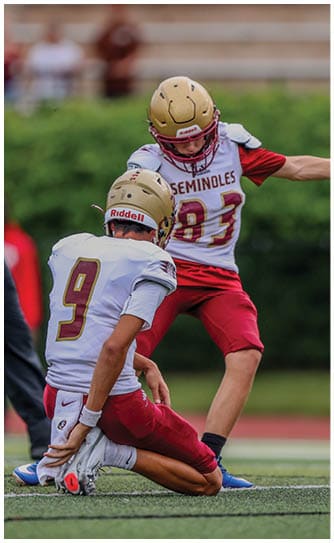 Two people wearing football helmets and Seminoles jerseys are engaged in a play on a field with a blurred green background. Description generated by AI