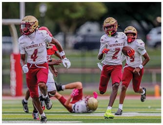 Adults wearing football uniforms with “Seminoles" on their jerseys are actively playing on a field, with one person running while holding a football and others in motion around them. Description generated by AI