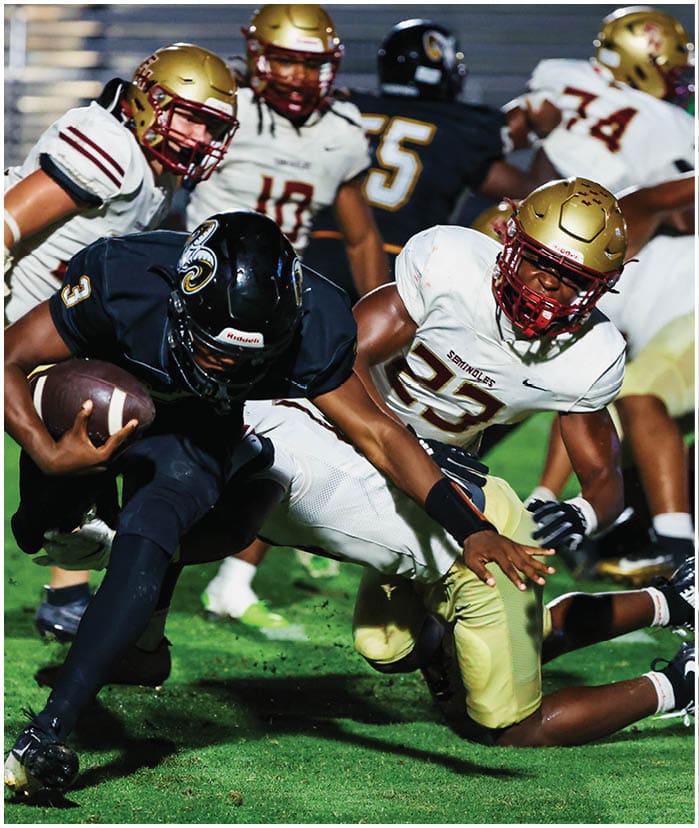 A person in a dark uniform is running with a football while being tackled by a person in a white and gold uniform during a football game. Description generated by AI