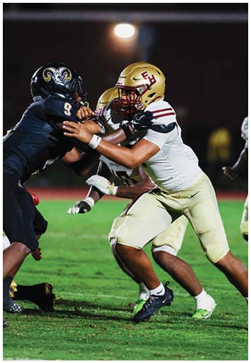 Two people wearing gold football helmets and white jerseys are engaged in a play on a dimly lit field at night. Description generated by AI