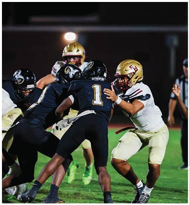 A person wearing a gold football helmet and uniform is seen behind several black helmets with a ram logo under bright stadium lights at night. Description generated by AI