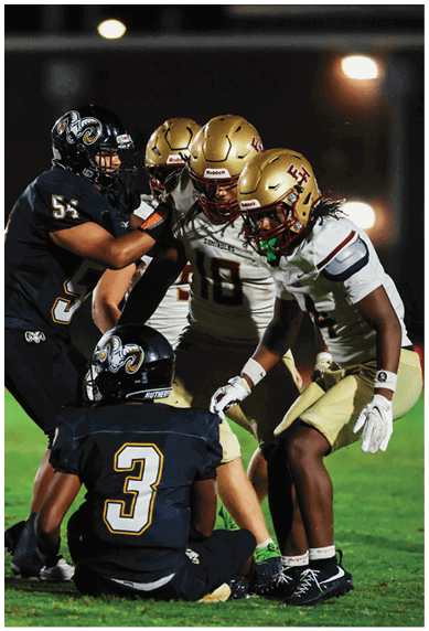 Four people wearing football helmets and uniforms are engaged in a close interaction on a dark field. Description generated by AI